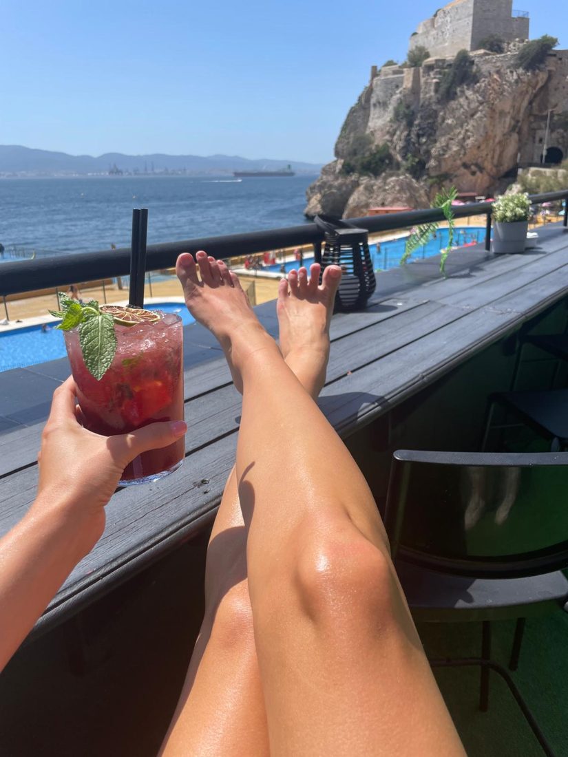 Bare legs stretched out on a rooftop bar with a cocktail and the sea behind